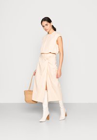 Top cropped bege e uma saia longa com detalhe torcido, acompanhados de botas brancas e uma bolsa de vime. Textura suave, design minimalista.