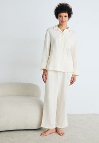 Lys beige, teksturert loungewear-sett bestående av en skjorte med knapper og lomme samt vide bukser, begge laget av mykt og pustende stoff.