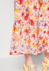 Midikleid mit floralem Muster in Rosa, Rot, Gelb und Lila, getragen mit gelben Plateau-Sandalen mit Blumenmuster.