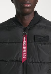 Svart vadderad bomberjacka med ribbad krage, zip-stängning och en röd tagg med texten "TA BORT INNAN FLYGNING" fäst vid dragkedjan.