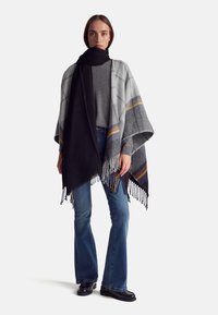 Poncho de cuadros gris con franjas amarillas y negras, con un dobladillo con flecos, combinado con una bufanda negra, usado sobre una blusa gris y unos vaqueros acampanados azules.