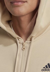 Parte inferior de la cara y cuello de una persona que lleva una sudadera con cremallera de Adidas en color beige, con capucha y tirador de cremallera visible.