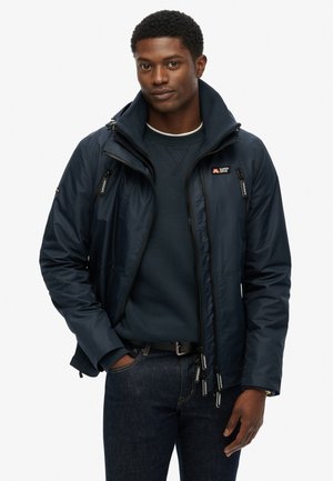Veste zippée bleu marine avec un col haut, dotée de poignets ajustables et d'un logo visible. Portée par-dessus un pull sombre et un jean.