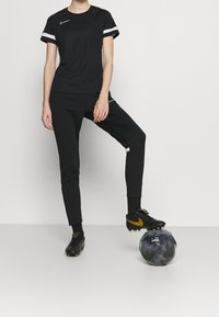 Schwarzes Sport-T-Shirt mit kurzen, weiß gestreiften Ärmeln, schwarze Jogginghosen und schwarze Turnschuhe mit goldenen Akzenten. Ball unter dem Fuß platziert.