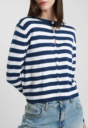 Donna che indossa un cardigan a righe orizzontali blu navy e bianche con bottoni, abbinato a jeans di jeans azzurri, su uno sfondo semplice.