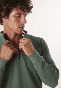 Hellgrüner Reißverschluss-Pullover mit gerippter Textur, ausgestattet mit einem Viertel-Reißverschluss-Kragen und einem kontrastierenden gestreiften Akzent im Inneren des Kragens.