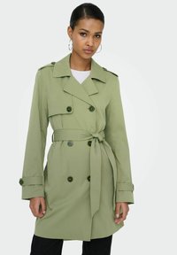 Femme portant un trench-coat ceinturé vert clair à boutons croisés et des boucles d'oreilles en forme d'anneau, debout devant un fond uni.