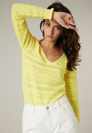 Femme aux longs cheveux foncés portant un pull à motifs jaune à col en V et un pantalon blanc, levant son bras gauche au-dessus de son front.