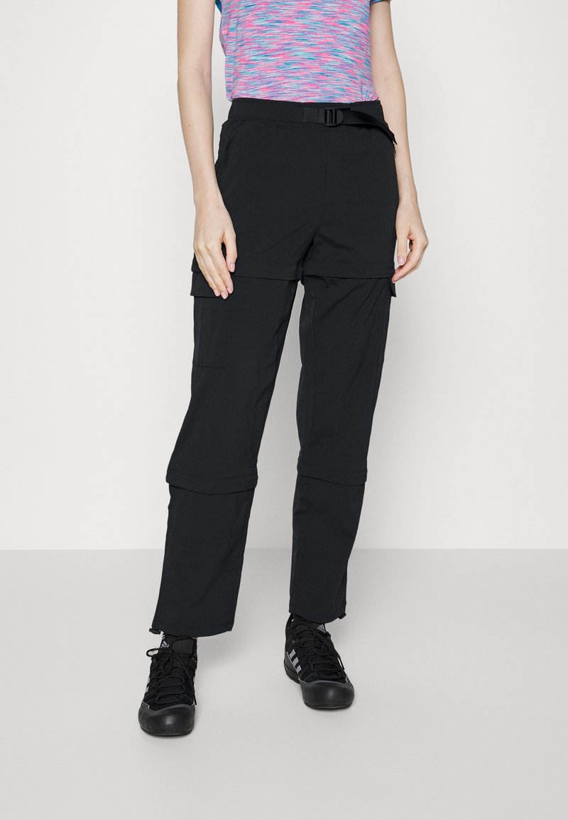 Personne portant un pantalon cargo noir convertible avec jambes zippées et des chaussures de sport noires, debout devant un fond blanc uni.