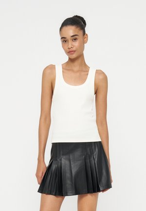 Femme aux cheveux attachés en chignon, portant un haut blanc sans manches et une jupe plissée en cuir noire, debout devant un fond clair uni.
