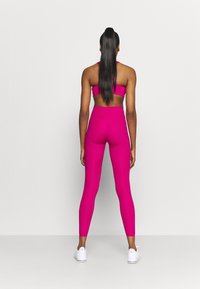 Nainen seisoo seinää vasten kirkkaan vaaleanpunaisessa urheiluoliivissa ja leggings-housuissa, valkoisissa lenkkareissa ja pitkät suorat hiukset poninhännällä.