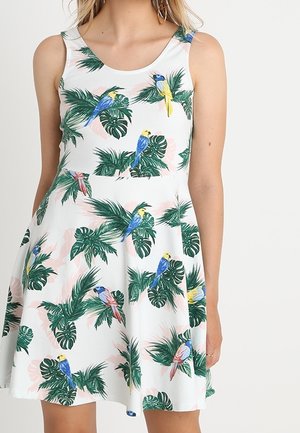 Mouwloze witte jurk met groene tropische bladeren en kleurrijke papegaaienprint, getailleerde taille, gedragen door een vrouw met een lichte huid.