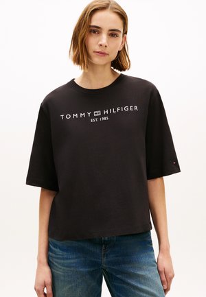 Giovane donna con capelli castani corti che indossa una maglietta nera Tommy Hilfiger e jeans blu, in piedi davanti a uno sfondo semplice.