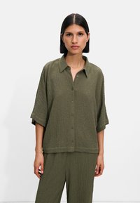Zielona oliwkowa koszula z guzikiem, z kołnierzykiem i krótkimi rękawami, o luźnym kroju i subtelnym, tkanym wzorze.