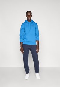 Μπλε φούτερ με κουκούλα, μπροστινή τσέπη και κορδόνια, συνδυασμένο με σκούρο μπλε φόρμα και λευκά αθλητικά παπούτσια· διαθέτει λογότυπο στο στήθος και το μανίκι.