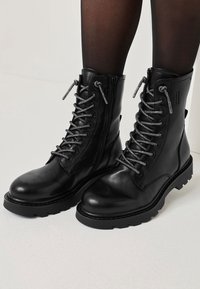 Bottes de cheville en cuir noir à texture lisse, bout rond, lacets gris et fermetures éclair latérales. Semelle robuste pour une meilleure adhérence.