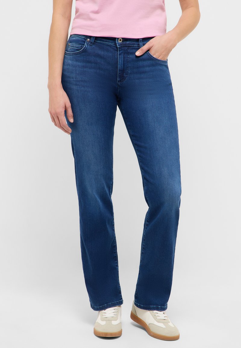 Blauwe spijkerbroek met rechte pijpen, voorzien van een voorzak, knoopsluiting en lichte vervaagde details. Draagt lichtgekleurde sneakers.