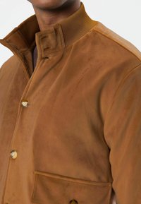 Brown suede jacket with a high collar, featuring a patch pocket and button closure. Soft texture with subtle sheen.