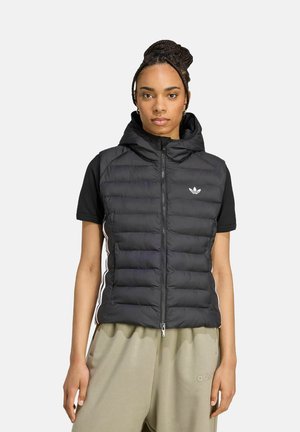 Frau mit geflochtenem Haar, die eine schwarze ärmellose gepolsterte Weste mit Kapuze über einem schwarzen kurzärmeligen Shirt und beigen Adidas-Hosen trägt.