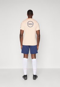 Light peach athletic shirt with "BARÇA" logo on the back. Worn with navy shorts, white knee socks, and black sports shoes.