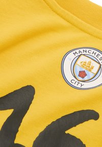 Gul bomulds T-shirt med et sort grafisk design og et rundt Manchester City-logo med skib- og rose-detaljer.