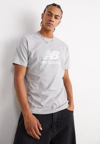 Pilkas medvilnės marškinėliai su balta "New Balance" logotipu, derinami su juodais laisvais kelniais. Modelis turi pynėmis susuktus plaukus ir kaklo papuošalą.