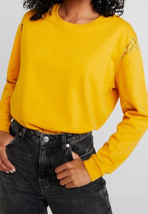 Kvinde med krøllet hår iført en gul bluse med lange ærmer og blonde detaljer på skuldrene samt højtaljede sorte denimjeans.