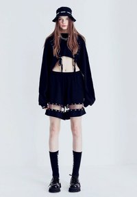 Suéter negro cortado con detalles de cordones, combinado con pantalones cortos negros que presentan acentos transparentes y una cadena. Llevado con un sombrero negro de tipo cubo y calcetines.