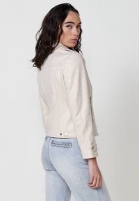 Jachetă bej, ajustată, cu guler, buzunare laterale și butoane cu capsenă. Material texturat și cusături contrastante. Purtată cu blugi de culoare albastru deschis.
