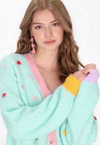 Junge Frau mit welligem Haar trägt eine pastellblaue Strickjacke mit pinken und roten Herzen, gelben und lavendelfarbenen Bündchen sowie pinken Herzohrhängern.