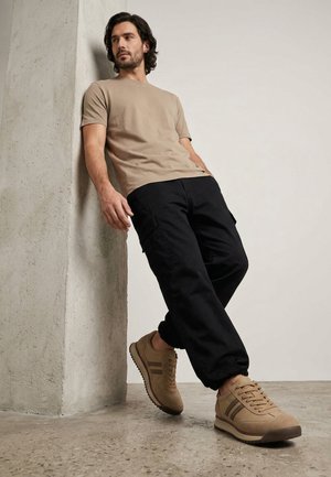 Mann mit dunklen Haaren, der ein beiges T-Shirt, schwarze Cargohosen und beige Sneakers trägt und an eine strukturierte Betonwand lehnt.