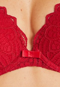 Bra rosso in pizzo caratterizzato da intricati motivi floreali, bordi scallop e un nastro di raso al centro, con un décolleté profondo.
