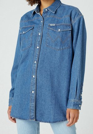 Woman wearing a long-sleeve blue denim shirt with snap buttons and two buttoned chest pockets, paired with light blue jeans.