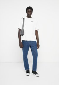 White cotton t-shirt with logo, blue denim jeans, gray crossbody bag, and black sneakers with white soles. Casual outfit on model.