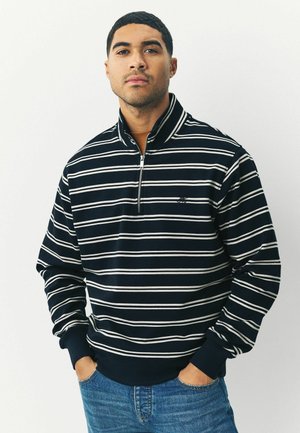 Navy striped pullover with a quarter-zip collar, featuring white horizontal stripes and ribbed cuffs, paired with blue denim jeans.