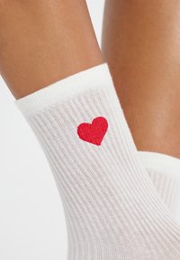 Calzini alla caviglia bianchi a coste con un piccolo ricamo a forma di cuore rosso sul lato. Materiale morbido, bordo elastico e texture liscia.