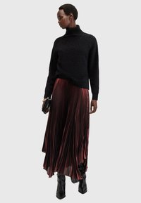 Maglione nero e texturizzato con collo alto, abbinato a una gonna plissettata borgogna. La modella indossa stivali neri lucidi fino al ginocchio e tiene una piccola borsa nera.