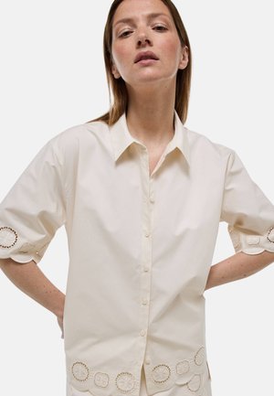 Femme portant une chemise beige à boutons avec des bords festonnés et une broderie circulaire découpée sur les manches et l'ourlet, debout les mains sur les hanches.