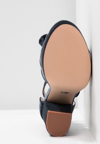 Sandale pour femme en daim noir avec talon bloc, semelle texturée visible et bride à la cheville, positionnée semelle vers le haut sur un fond blanc.
