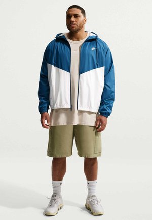 Mężczyzna stojący w niebiesko-białej kurtce Nike, beżowej oversize koszulce, oliwkowych spodenkach typu cargo, białych skarpetkach Nike i białych tenisówkach.