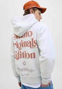 Sweatshirt à capuche gris avec un grand texte "Édition Origine Studio" en couleur rouille au dos, présentant un ourlet et des poignets côtelés.