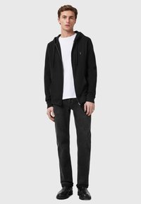 Felpa nera con zip e tasche frontali, indossata sopra una maglietta bianca, abbinata a jeans grigio scuro e scarpe nere. Design semplice e informale.