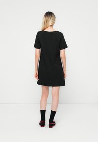 Black short-sleeve dress with a loose fit, knee-length hem, and a scoop back. Worn with black socks and red bow-accented shoes.