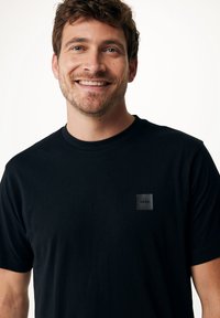Černé tričko s krátkým rukávem vyrobené z měkké látky, s čtvercovou černou nášivkou na levé hrudi a klasickým kulatým výstřihem.