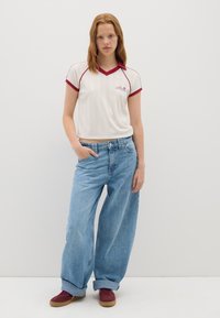 Witte korte mouwen shirt met een rode kraag en manchetten, gecombineerd met lichtblauwe baggy jeans. Model draagt bordeauxrode sneakers. Eenvoudig ontwerp en ontspannen pasvorm.