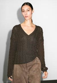 Modella che indossa un maglione lavorato a maglia marrone scuro con scollo a V e maniche ampie, abbinato a pantaloni scamosciati marrone chiaro, si trova davanti a uno sfondo semplice.
