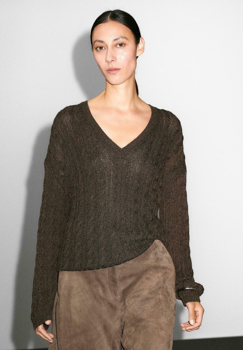 Modella che indossa un maglione lavorato a maglia marrone scuro con scollo a V e maniche ampie, abbinato a pantaloni scamosciati marrone chiaro, si trova davanti a uno sfondo semplice.