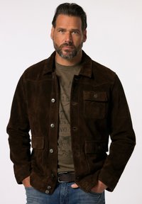 Chaqueta de ante marrón con botones al frente, dos bolsillos en el pecho y un collar. Llevada sobre una camiseta gráfica gris y combinada con jeans de mezclilla.