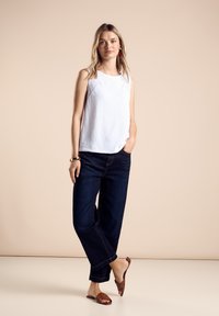 Witte mouwloze top van geweven stof met gaatjesdetails, gecombineerd met donkerblauwe wijde jeans en bruine platte sandalen. Neutrale achtergrond.