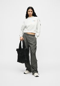 Νεαρή γυναίκα με μακριά μαύρα μαλλιά, φοράει λευκό κοντό φούτερ και γκρι παντελόνι cargo, κρατώντας μια μαύρη τσάντα ώμου, στέκεται μπροστά σε απλό φόντο.